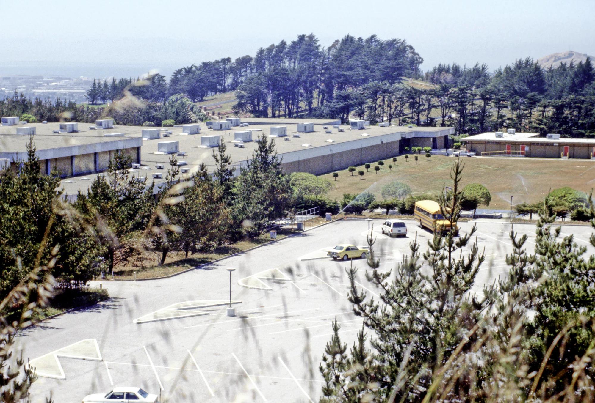 An elevated mid-1980s view of school buildings, a lawn and a parking lot with a yellow school bus below wooded hills
