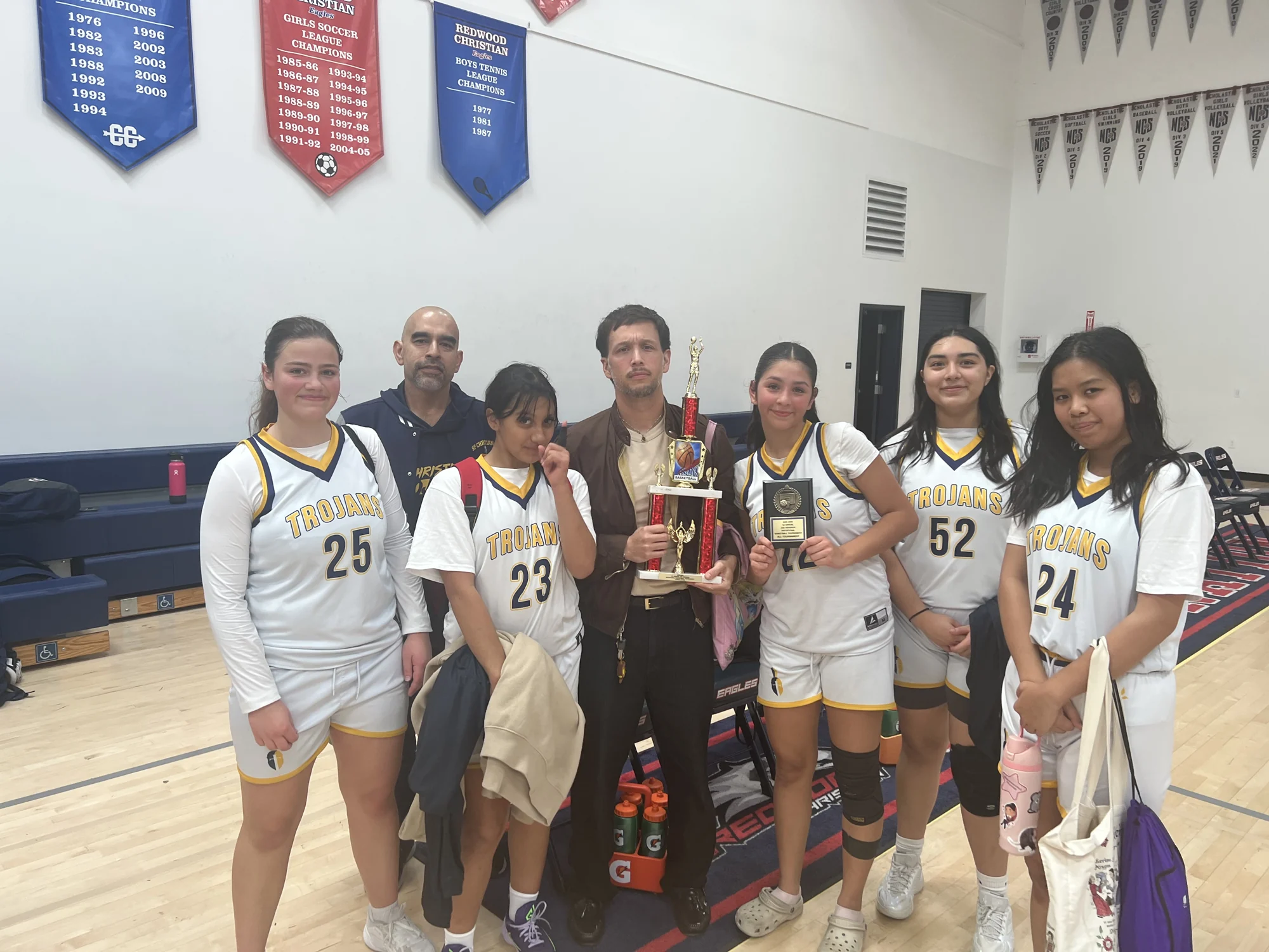 Trojans basketball players and coaches gathered around a trophy in the gym