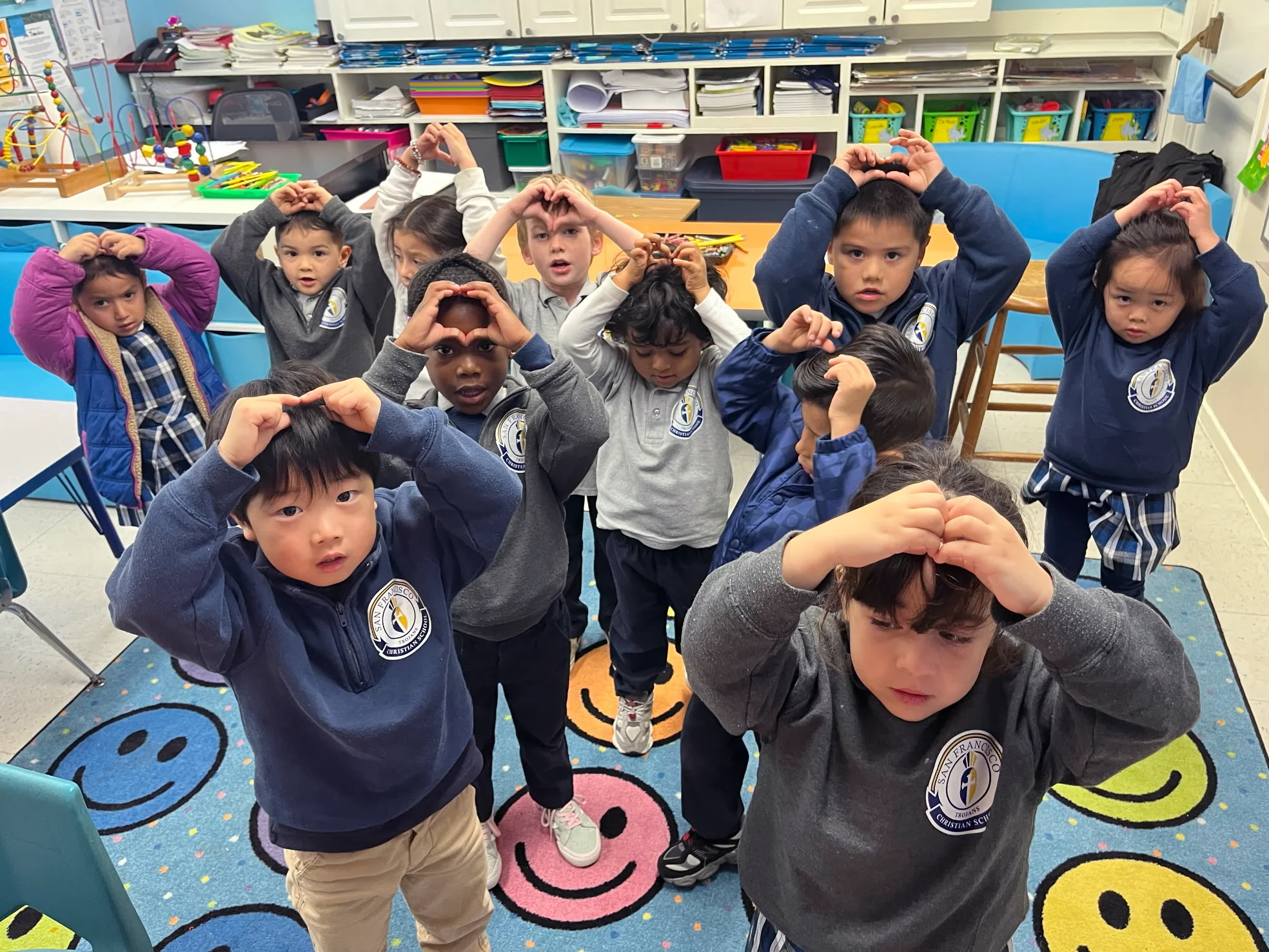 A kindergarten class making heart hands together on the smiley-face rug