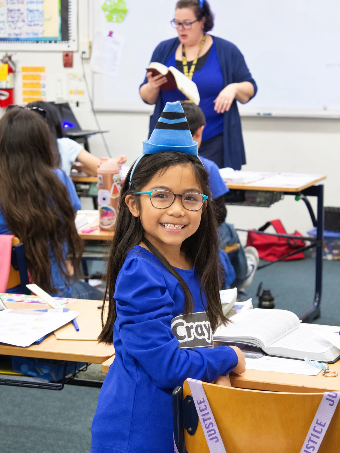 A girl in a blue crayon costume smiling in class while the teacher reads aloud behind her