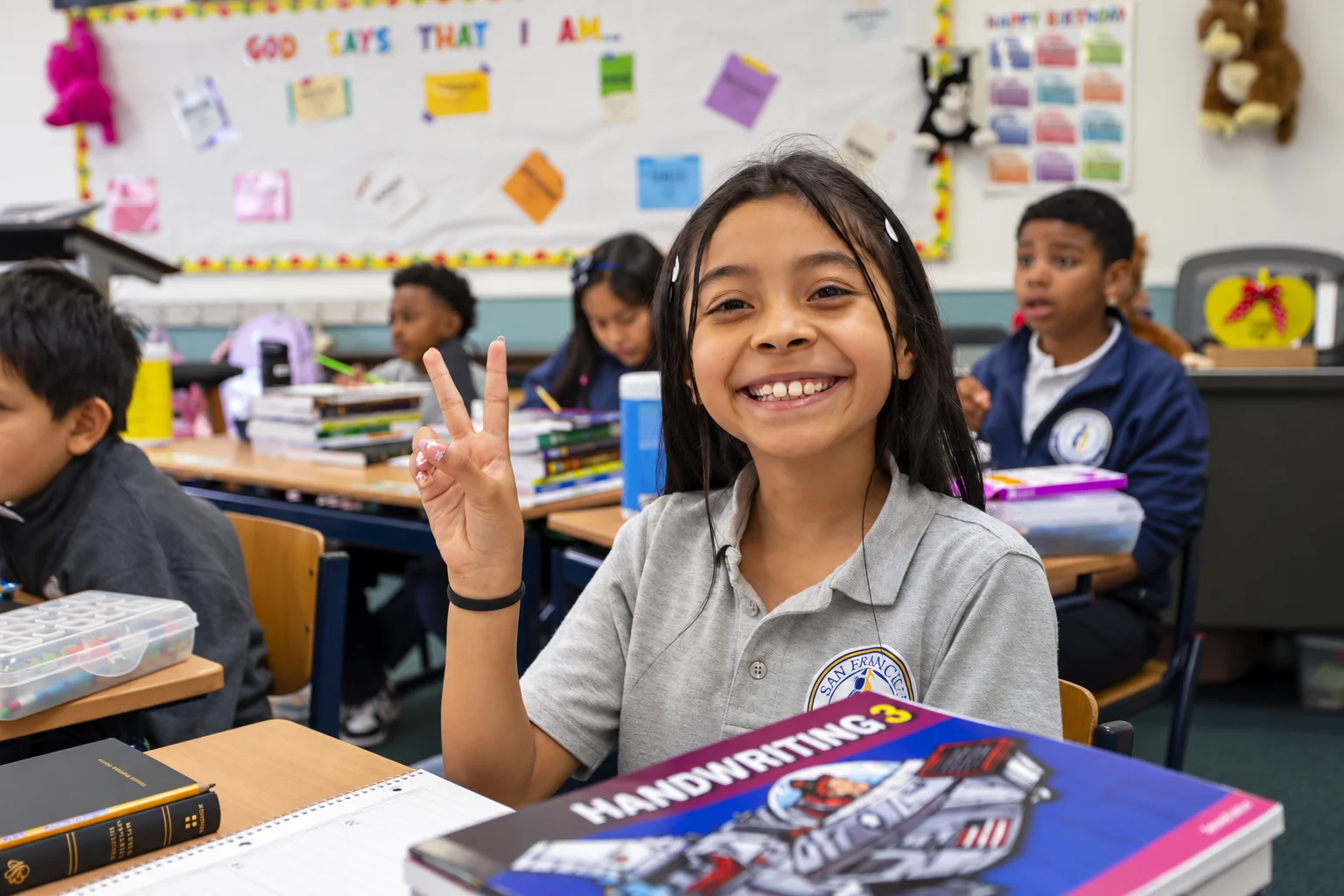 A girl flashing a peace sign over her handwriting workbook, classmates at their desks behind her