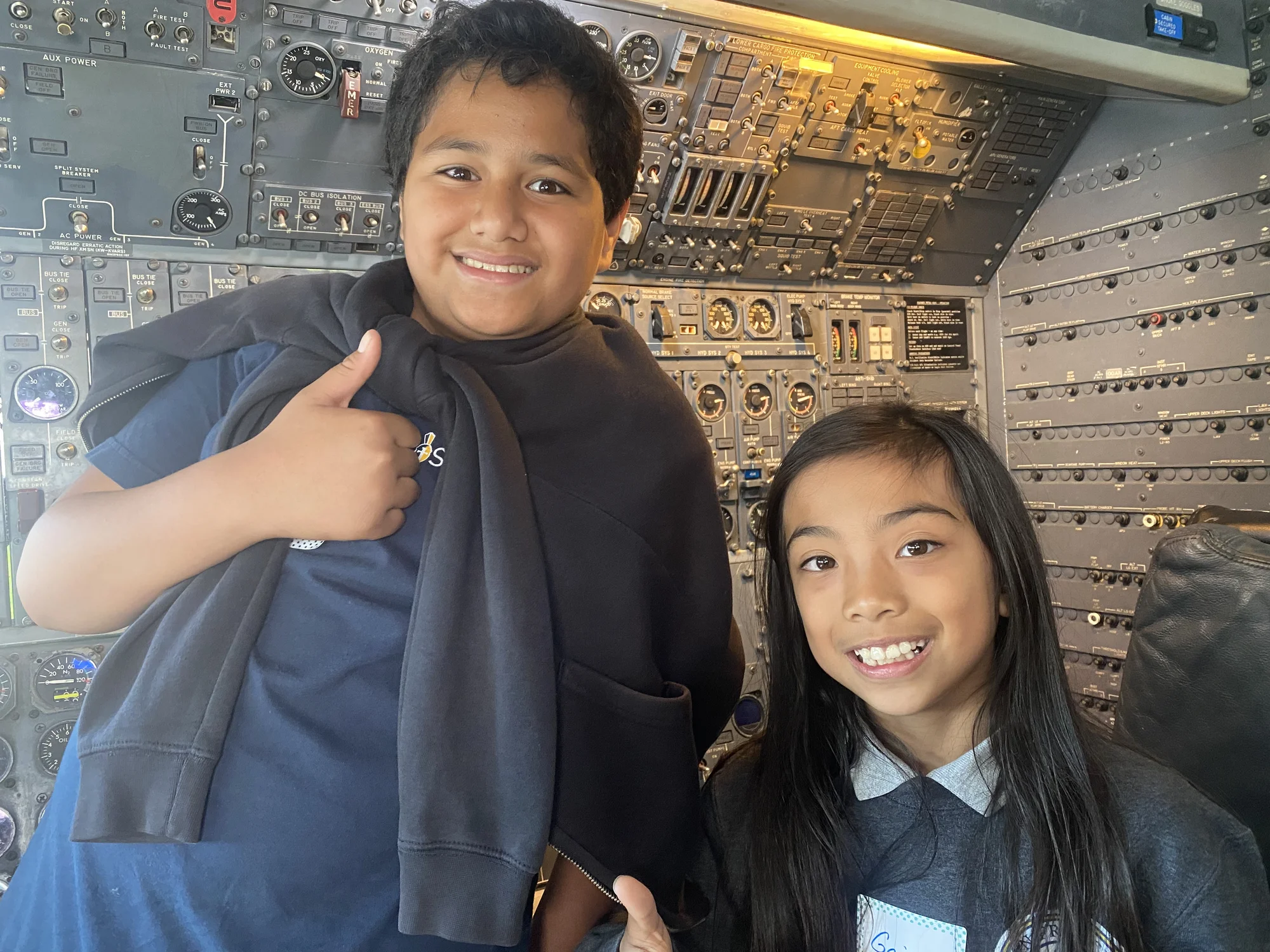 Two students grinning in an aircraft cockpit on a field trip, one giving a thumbs-up