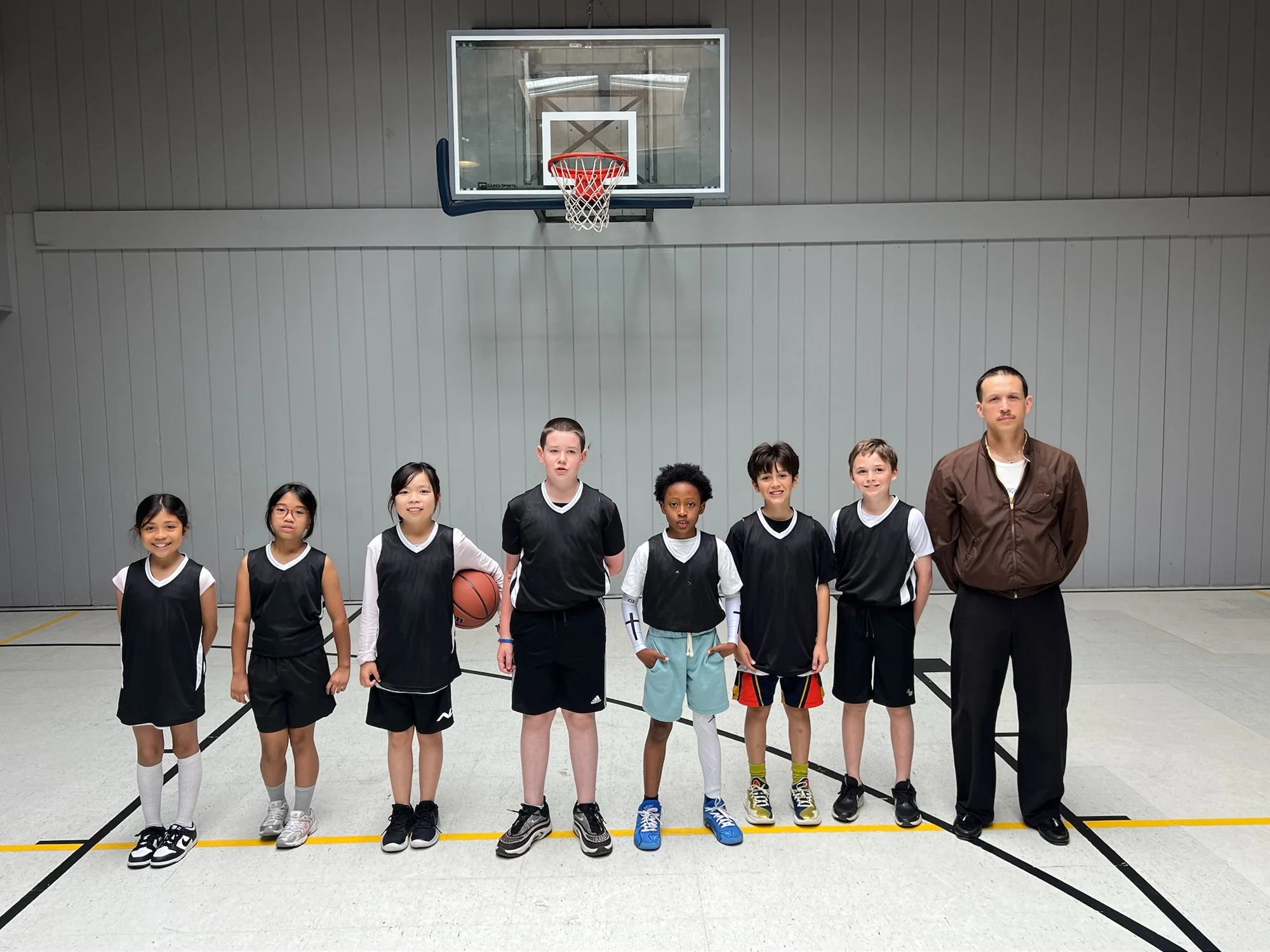 Elementary basketball players in black jerseys and their coach beneath the gym hoop