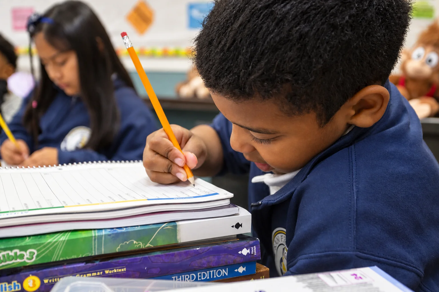 A boy bent close over his notebook, pencil in hand, at a stack of classroom workbooks