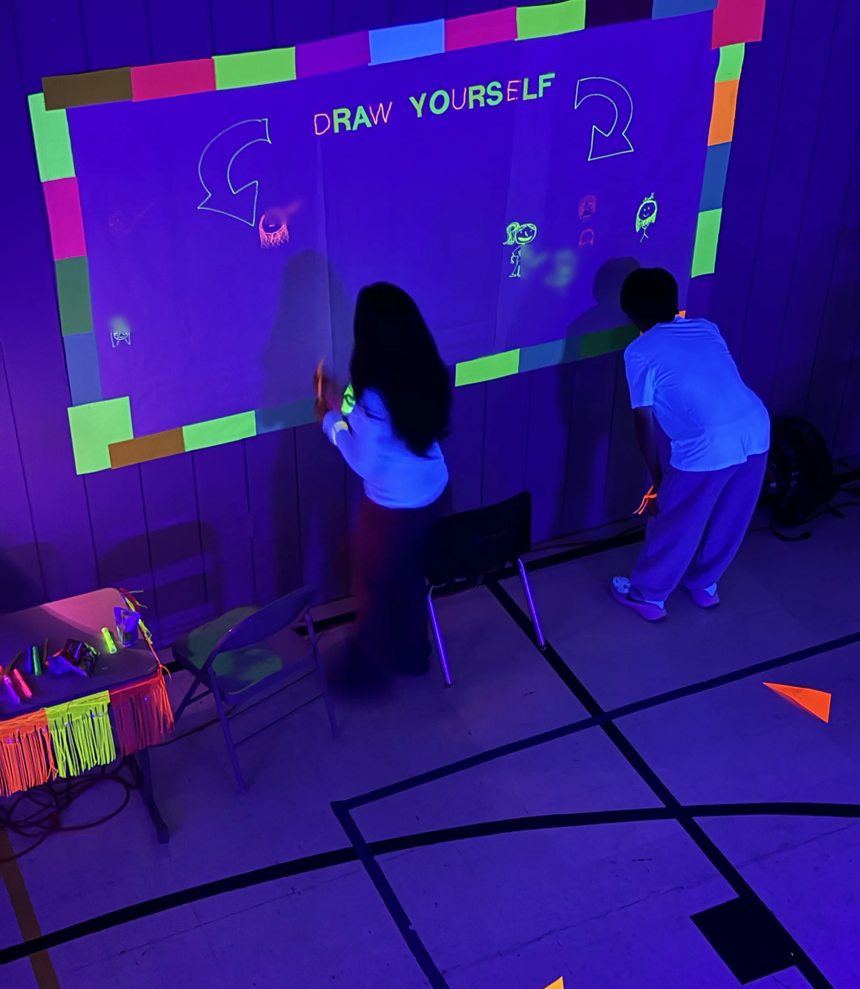 Two students adding their drawings to a glowing Draw Yourself wall at Glow Night