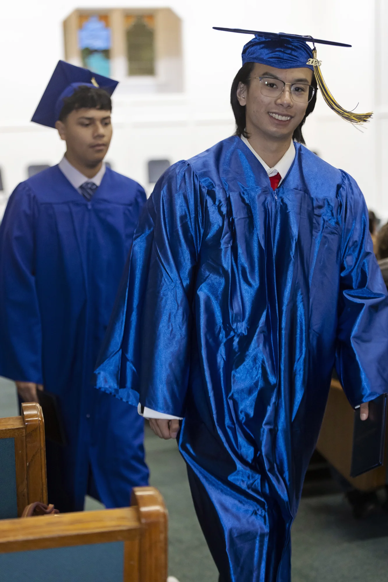 A smiling high school graduate in a blue cap and gown walks down the church aisle with classmates behind