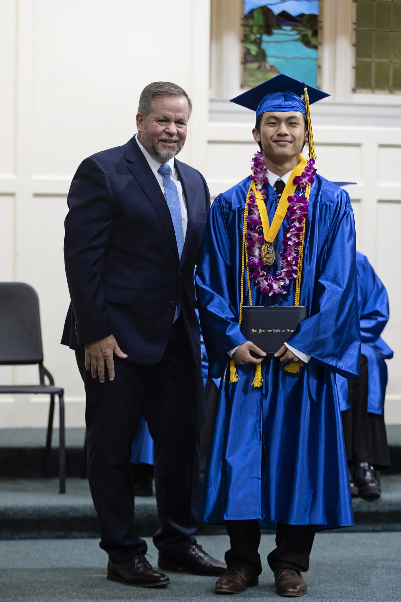 A high school graduate wearing a purple flower lei poses beside a school leader with a closed diploma folder