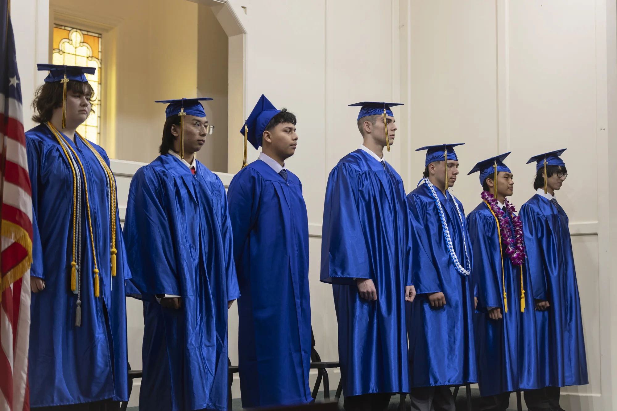 Seven high school graduates in blue caps and gowns stand together on the church stage