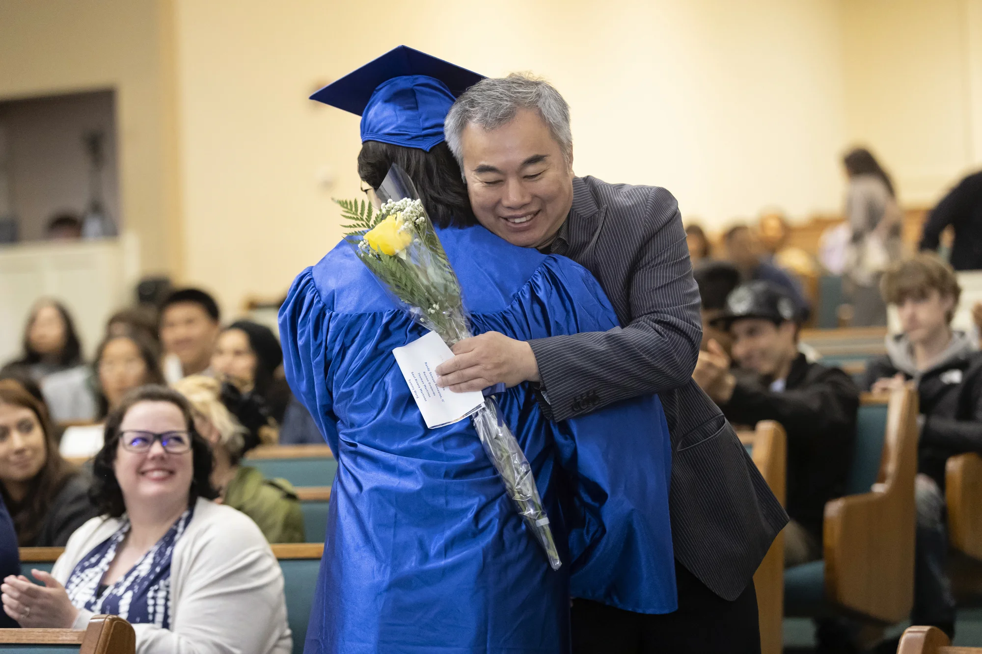 An attendee holding a wrapped yellow rose embraces a high school graduate in a blue cap and gown