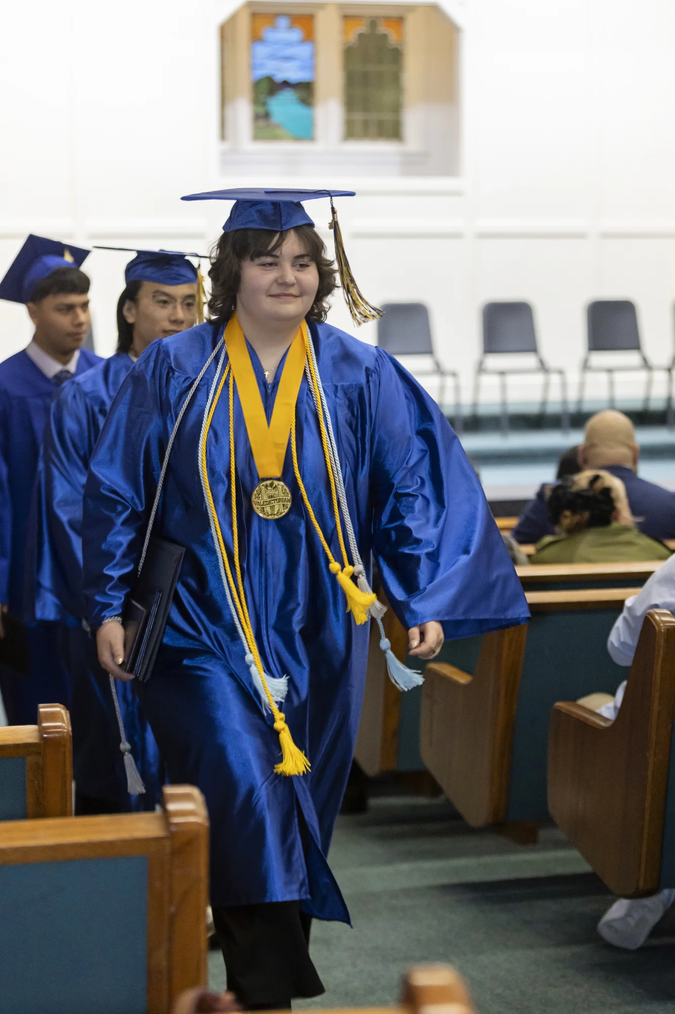 A high school graduate wearing gold cords leads classmates down the church aisle