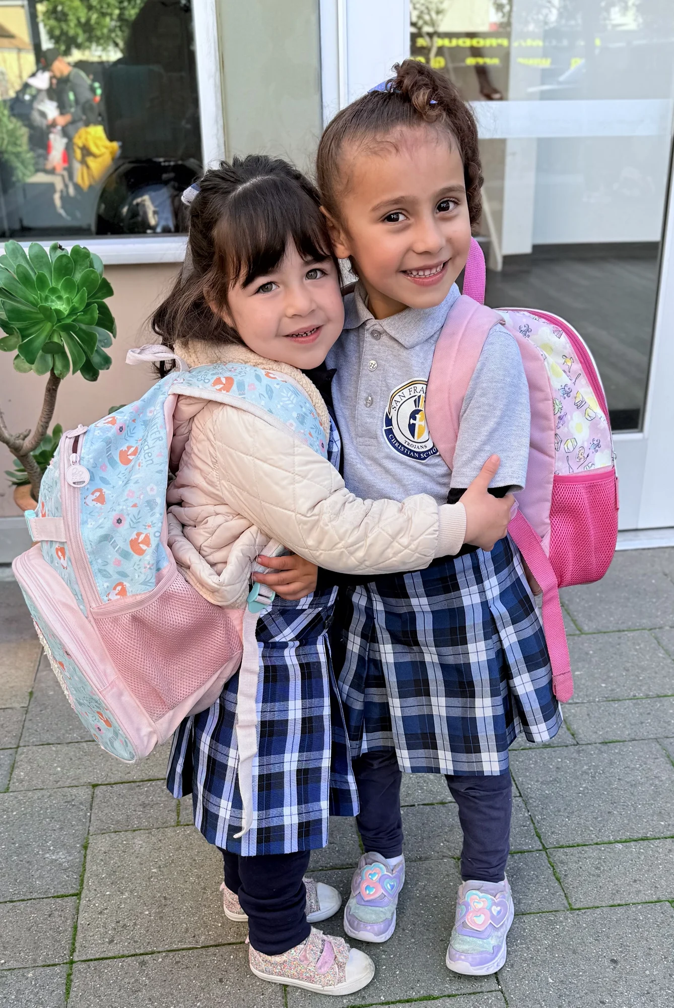 Two young students in plaid uniform skirts hugging with their backpacks on outside school