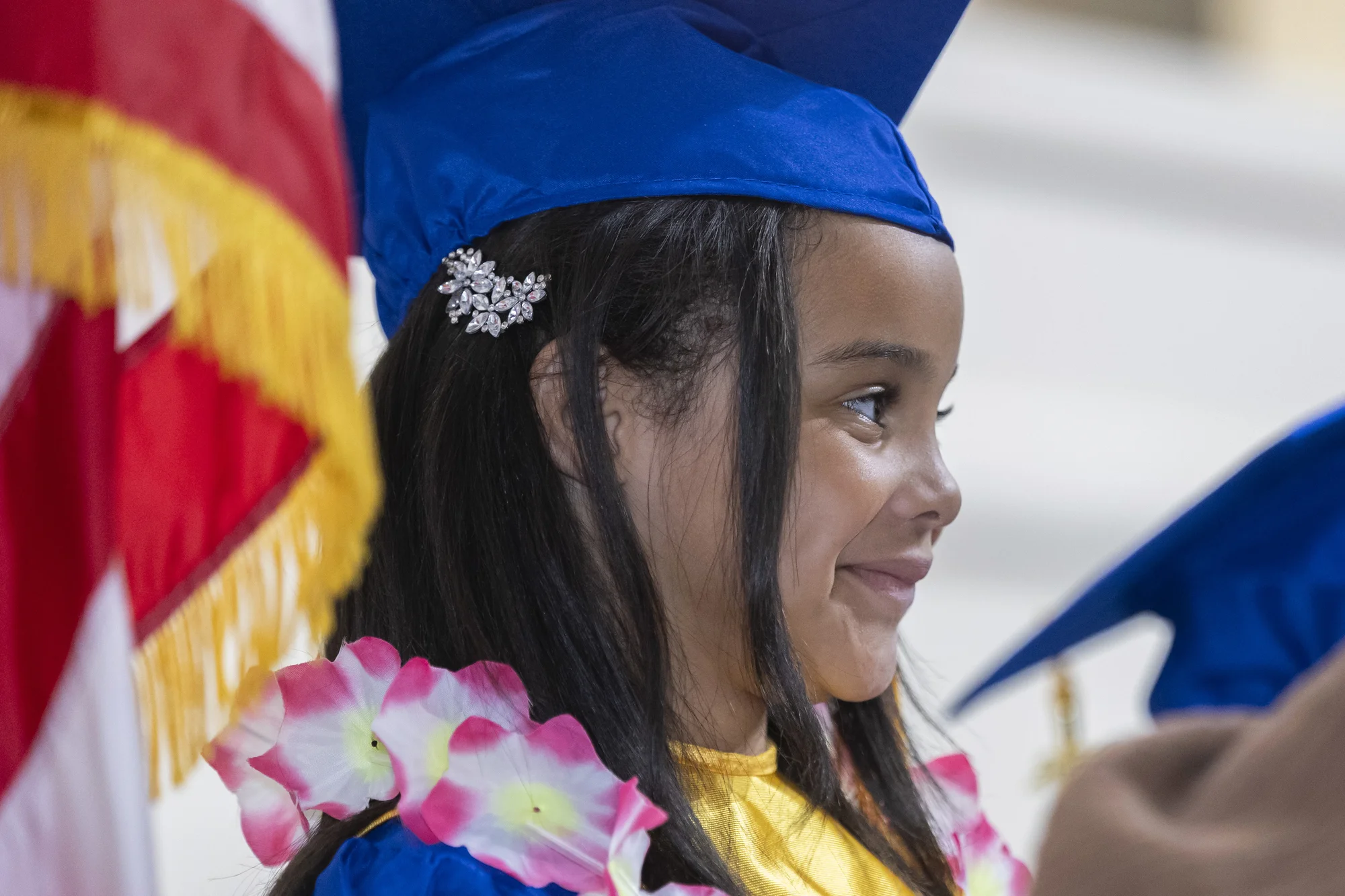 A kindergarten graduate in a blue cap and pink-and-white flower lei smiles in profile beside a flag