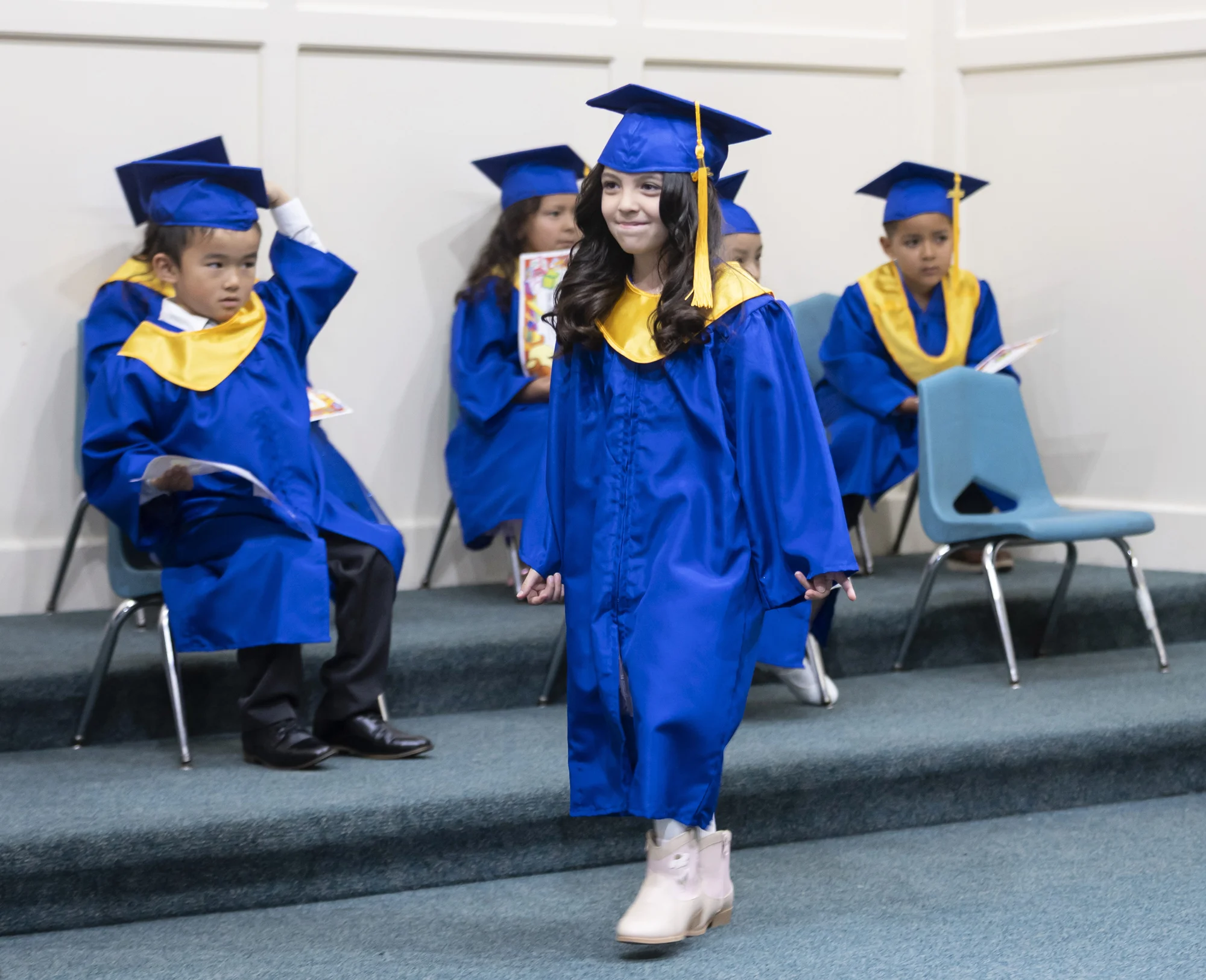 A kindergarten graduate in a blue cap and gown walks forward as classmates sit behind