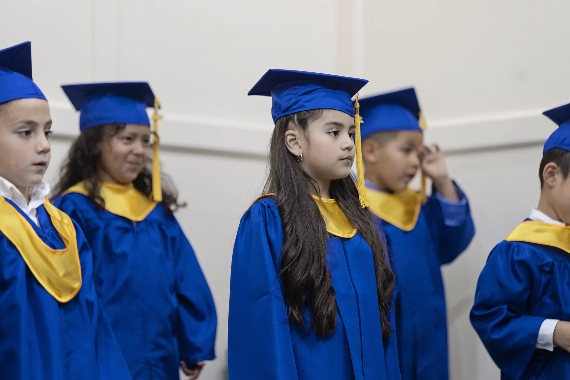 Children in blue graduation caps and gowns with gold collars stand together onstage