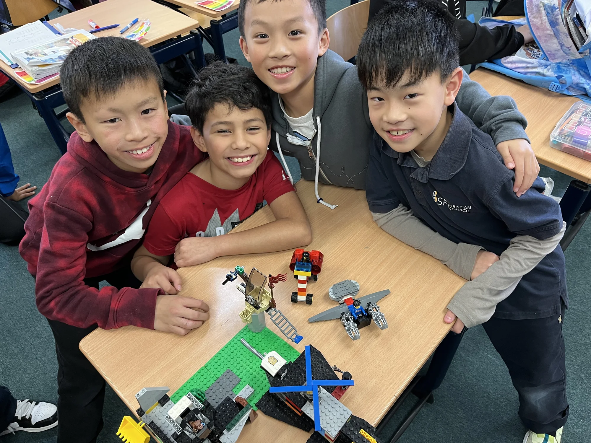Four grinning students leaning over the Lego builds on their classroom desk