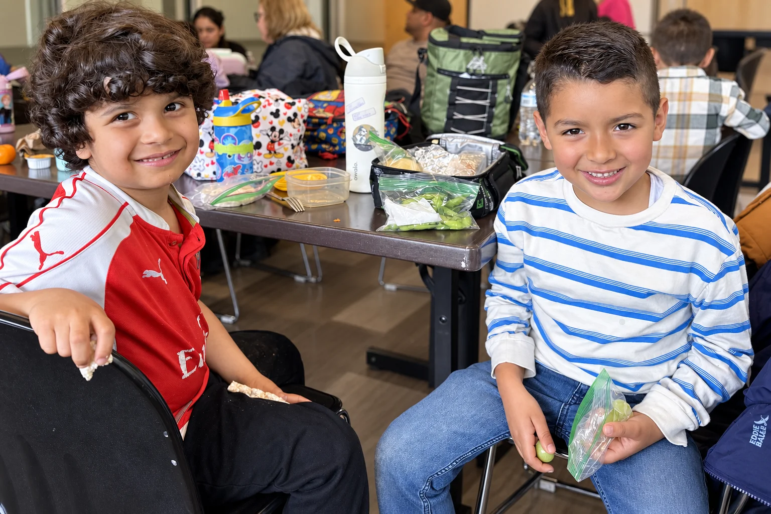 Two young students smiling at the lunch table with their lunches spread out
