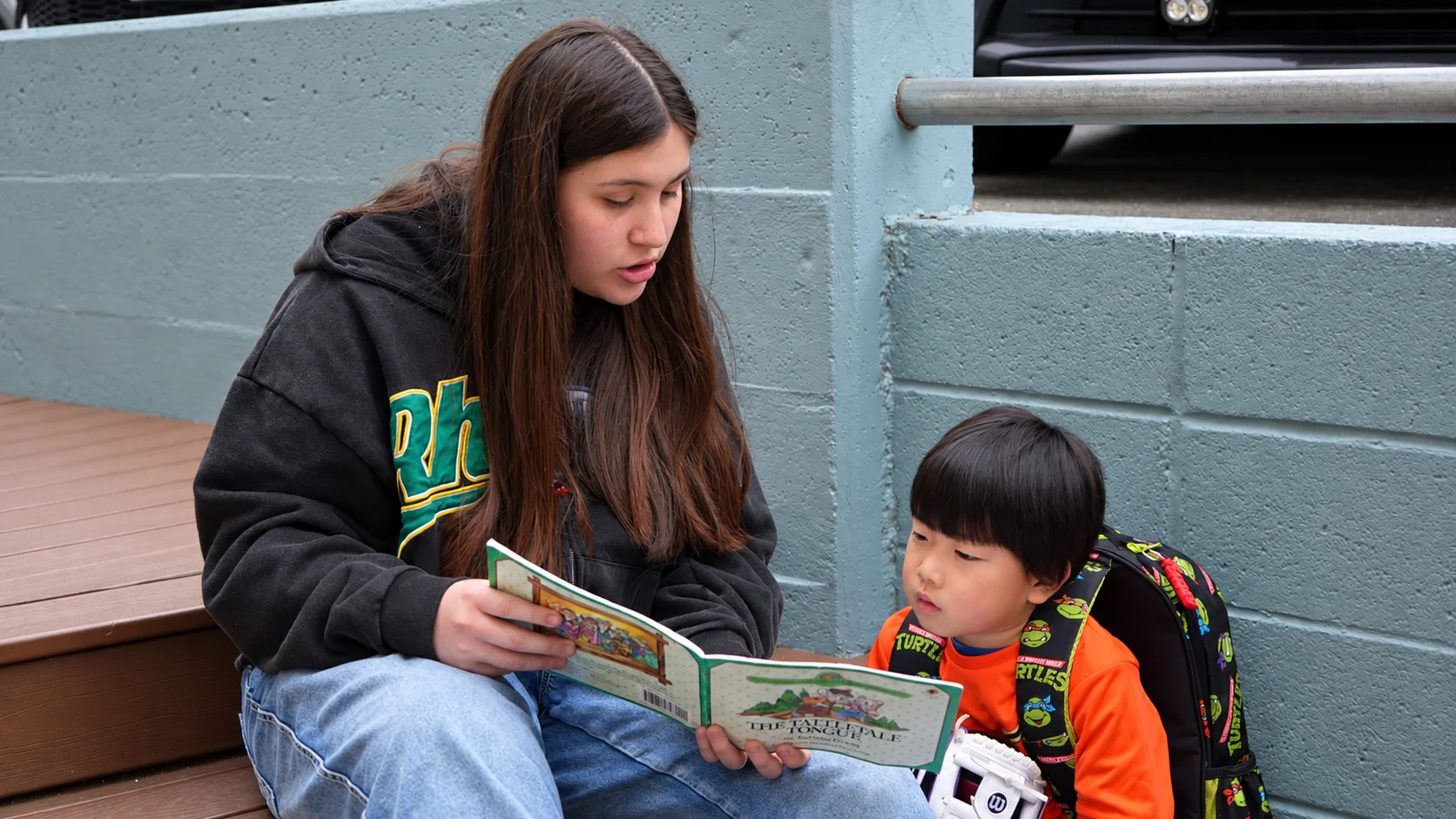 An older student reading a picture book aloud with a young boy on the outdoor steps
