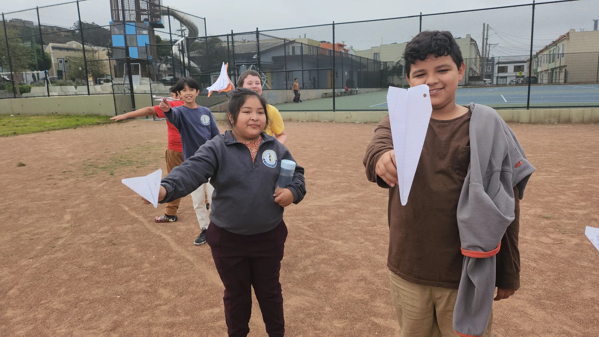 A boy and girl grinning with paper airplanes as their class lines up behind them