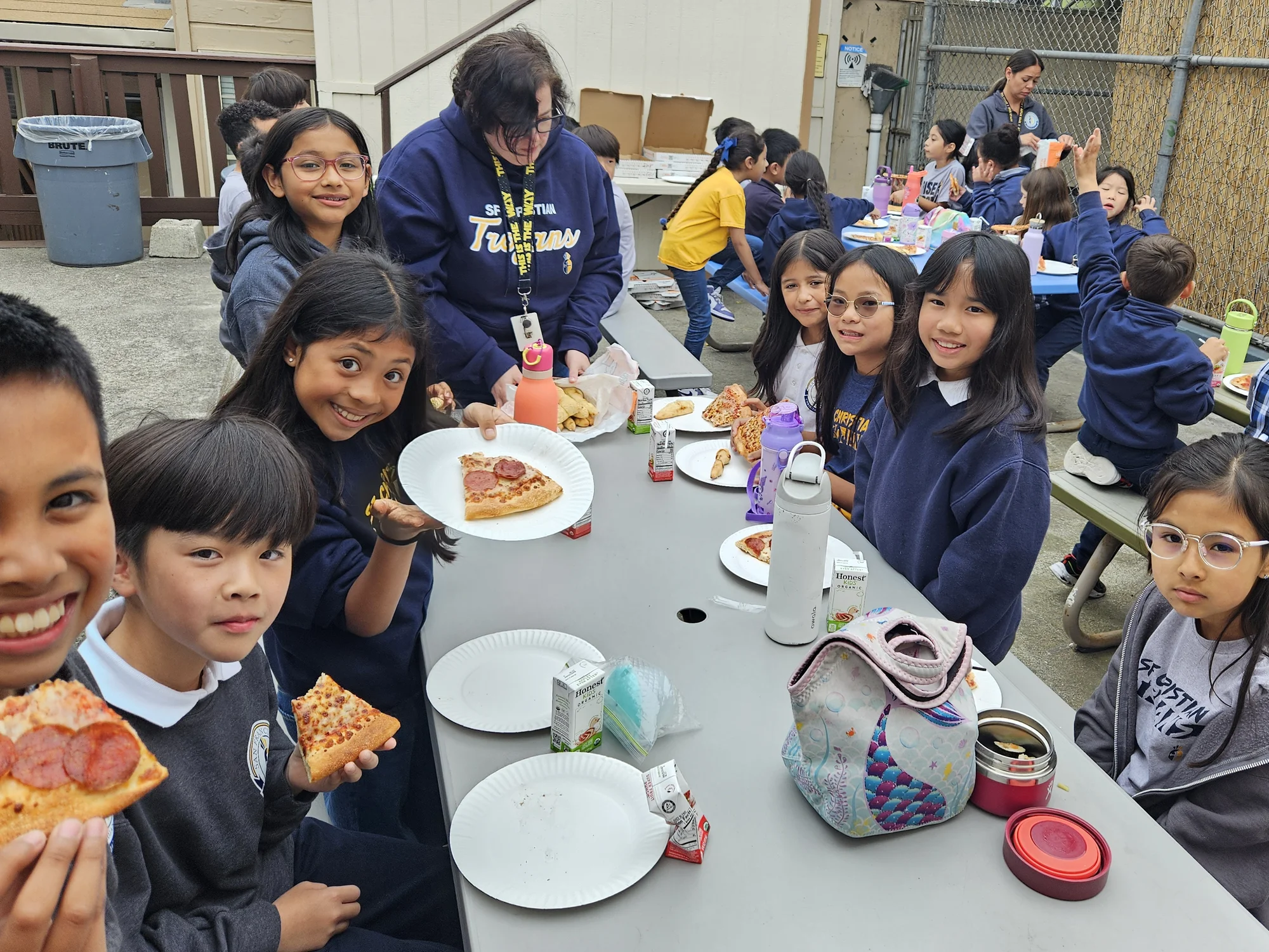 Students at a long outdoor table enjoying pizza together with staff