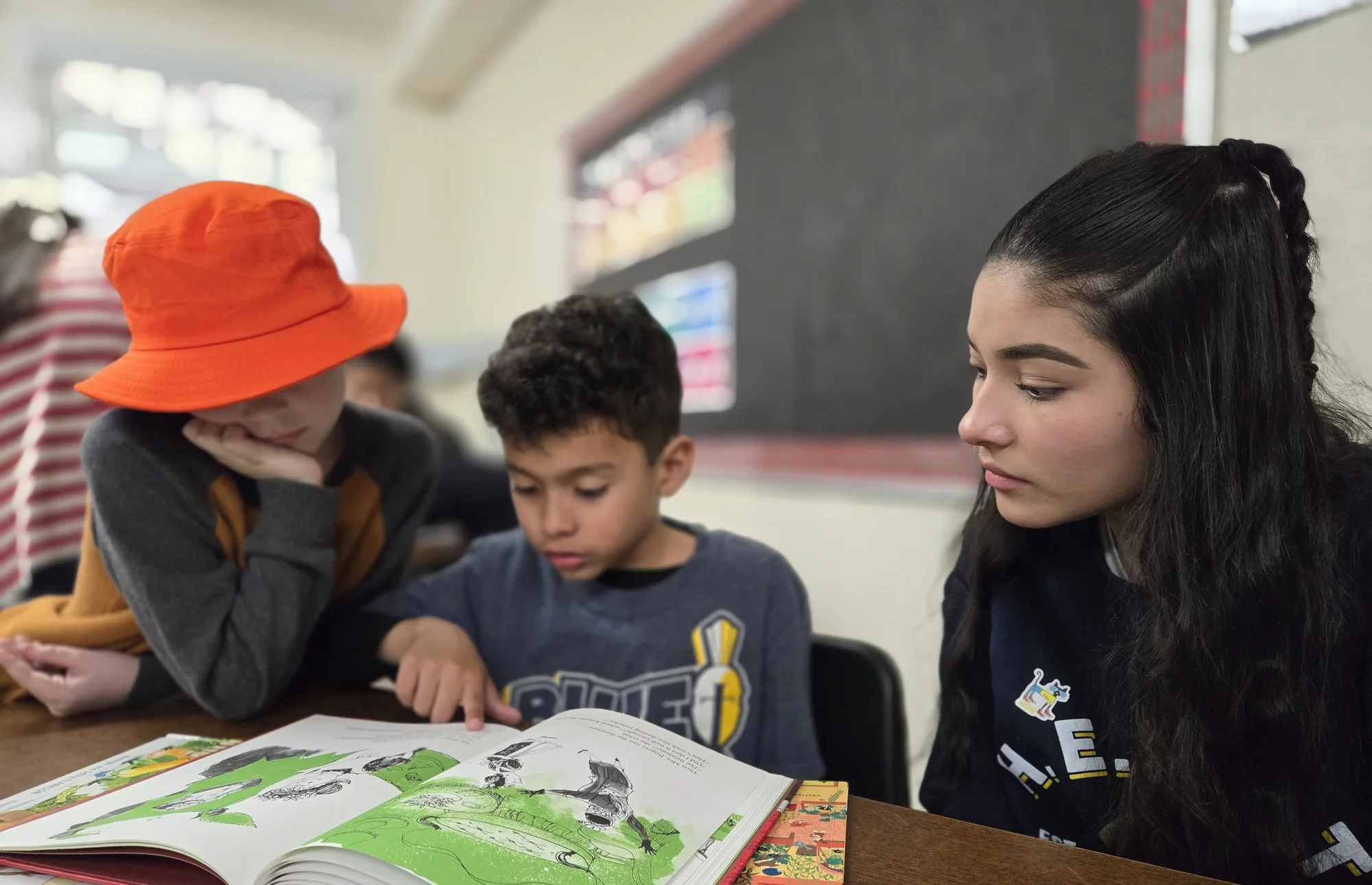 Three students reading a large picture book together at a classroom table