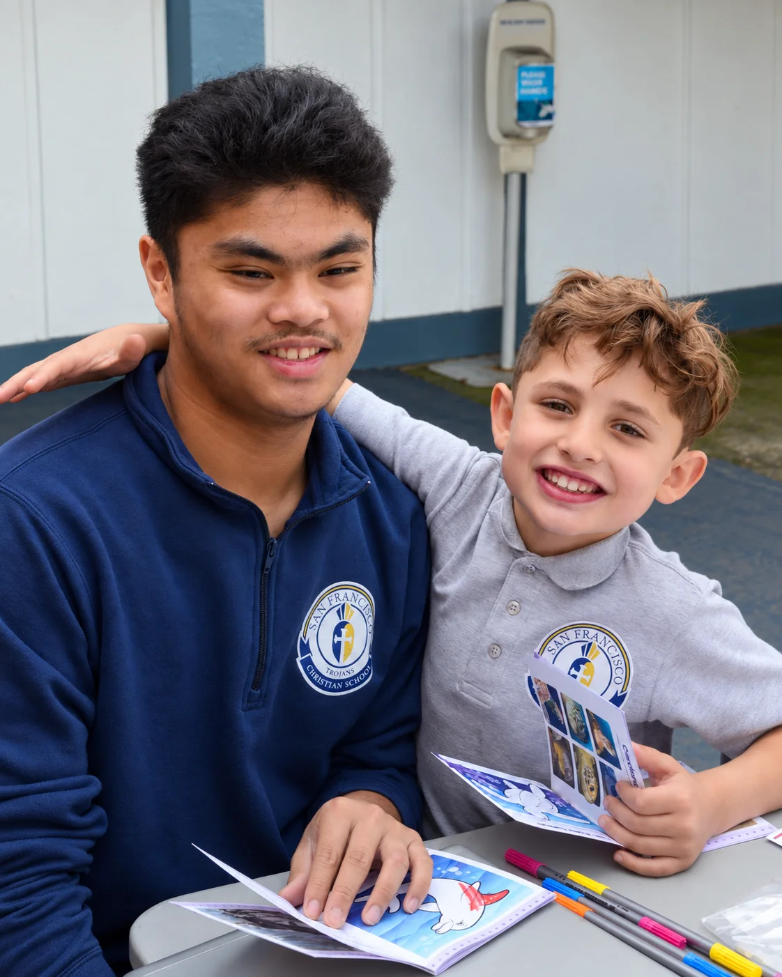 A high schooler with his arm around a young student, both smiling over their activity books