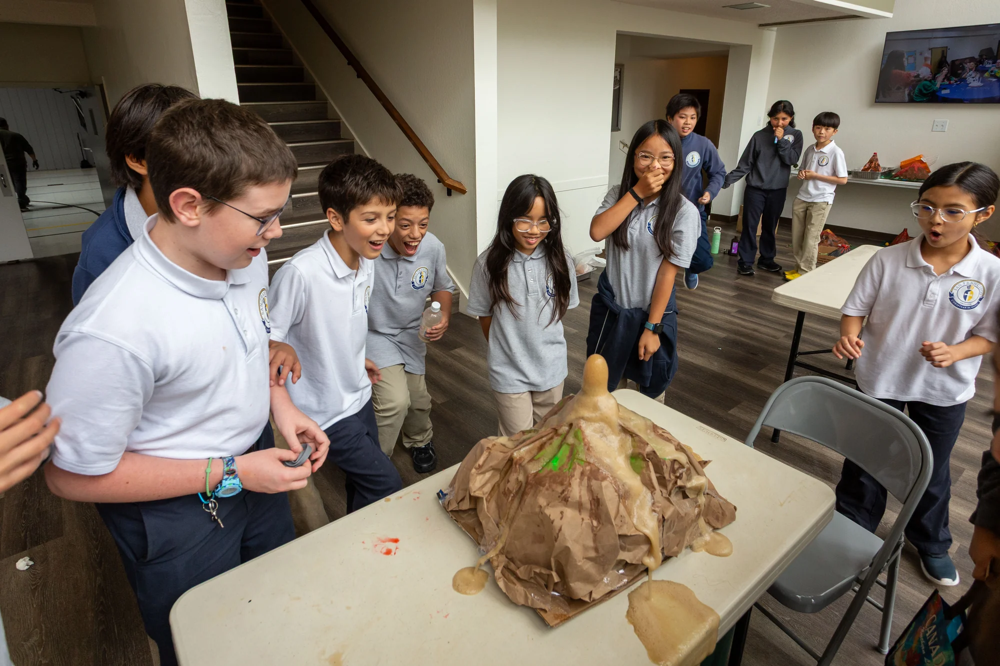 Sixth graders watch foam overflow from a large paper volcano on a classroom table