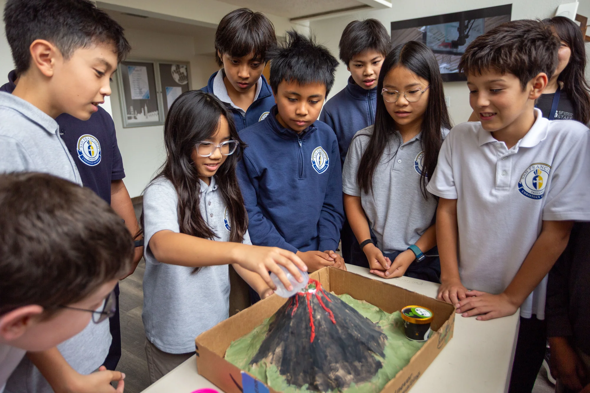 A sixth-grade student pours liquid into a model volcano while classmates gather around to watch