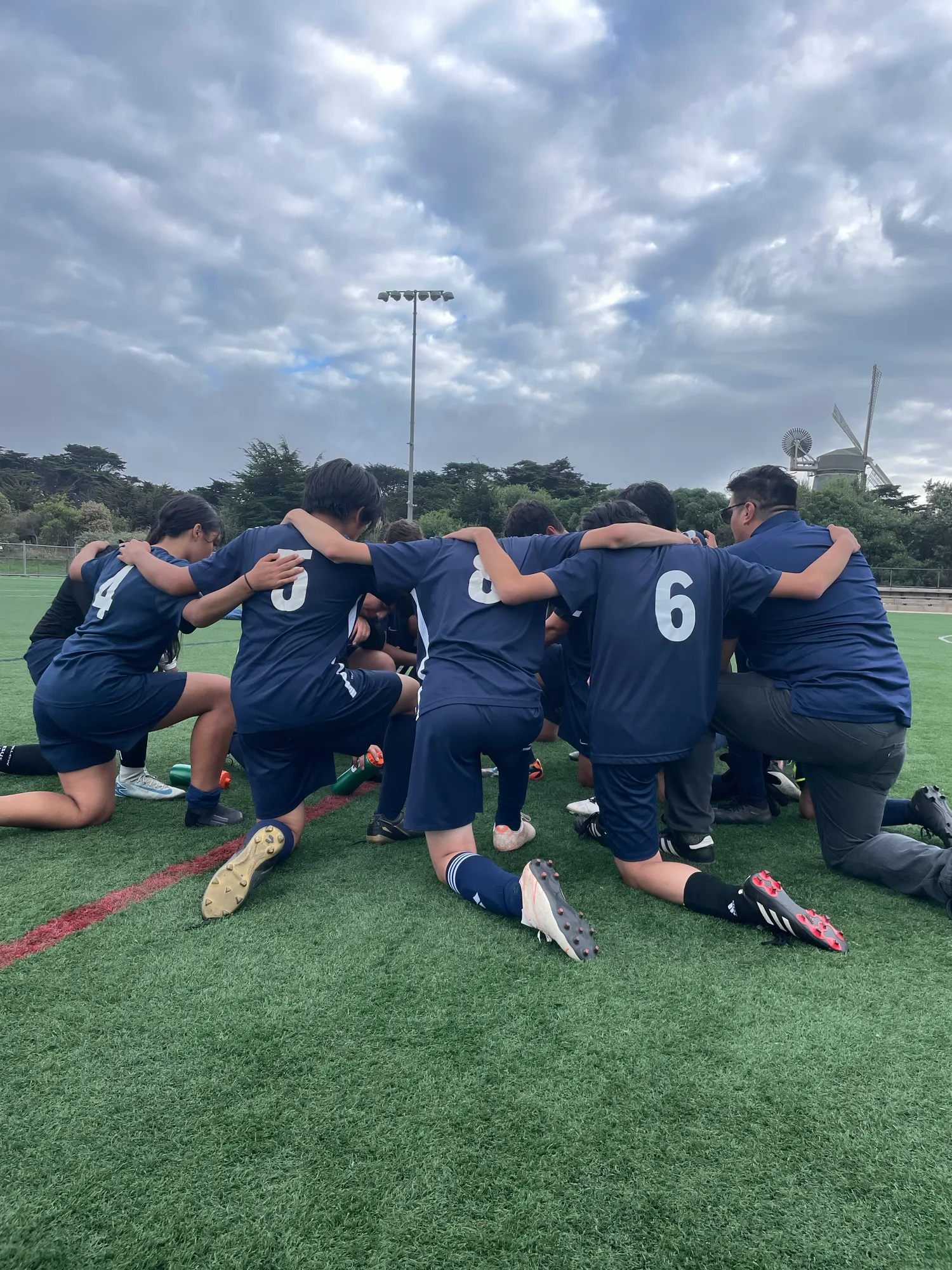 The soccer team kneeling in a huddle with their coach, arms around shoulders, the Golden Gate Park windmill behind