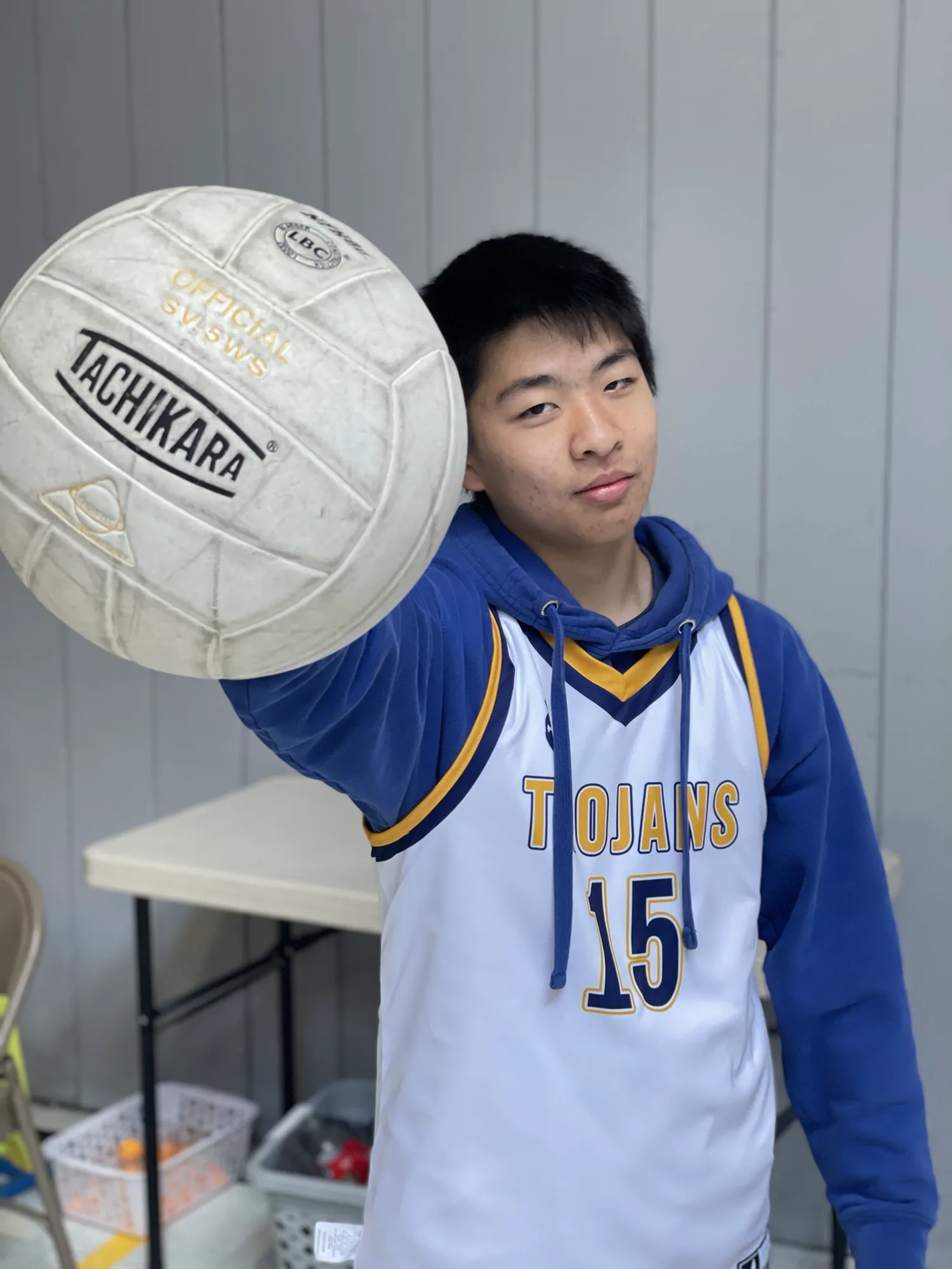 A high school volleyball player in a white Trojans jersey holding a volleyball out toward the camera