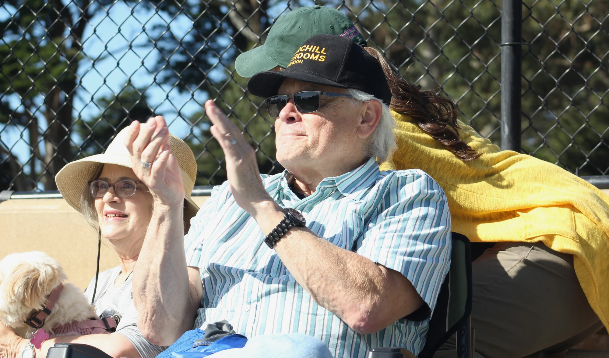 Parents applauding from the sideline at a soccer match, a small dog on a lap beside them