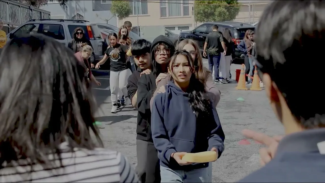 Students passing a foam baton in a relay race on the blacktop while classmates wait their turn