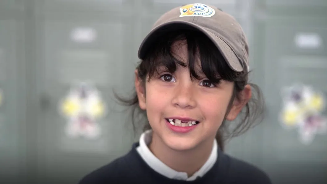 An elementary student in a school sweatshirt and cap smiling in front of the hallway lockers