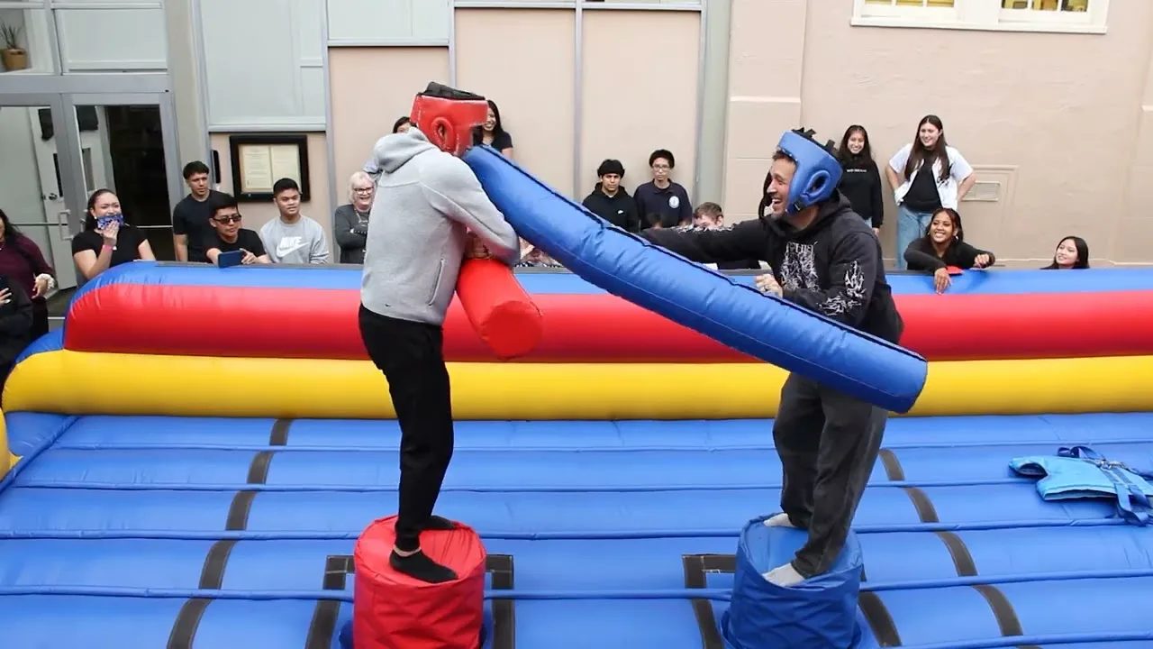 Two competitors in padded headgear balancing on pedestals in an inflatable jousting ring while students watch