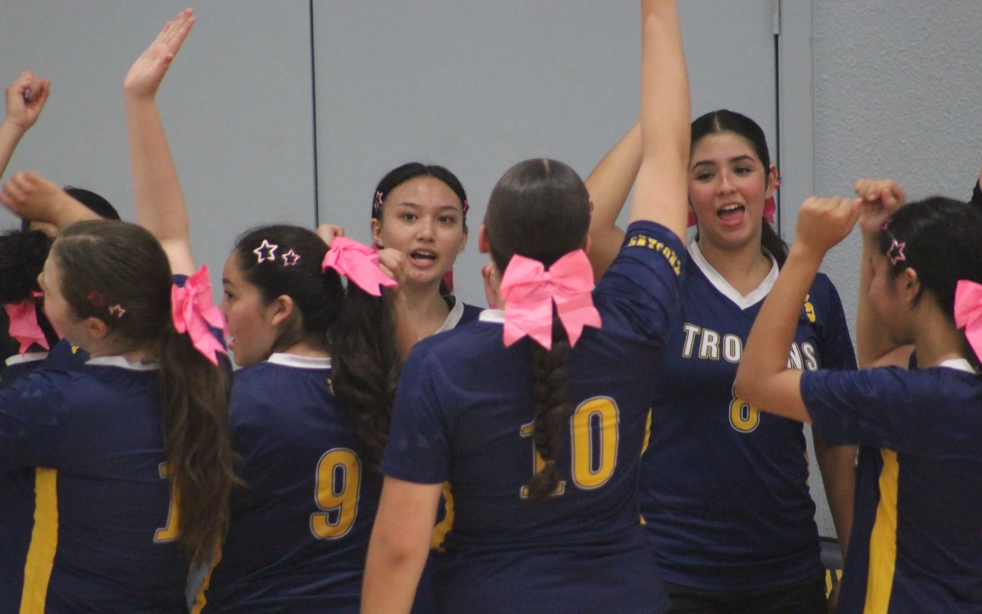 Trojans volleyball teammates in pink hair bows raising their arms together in a huddle